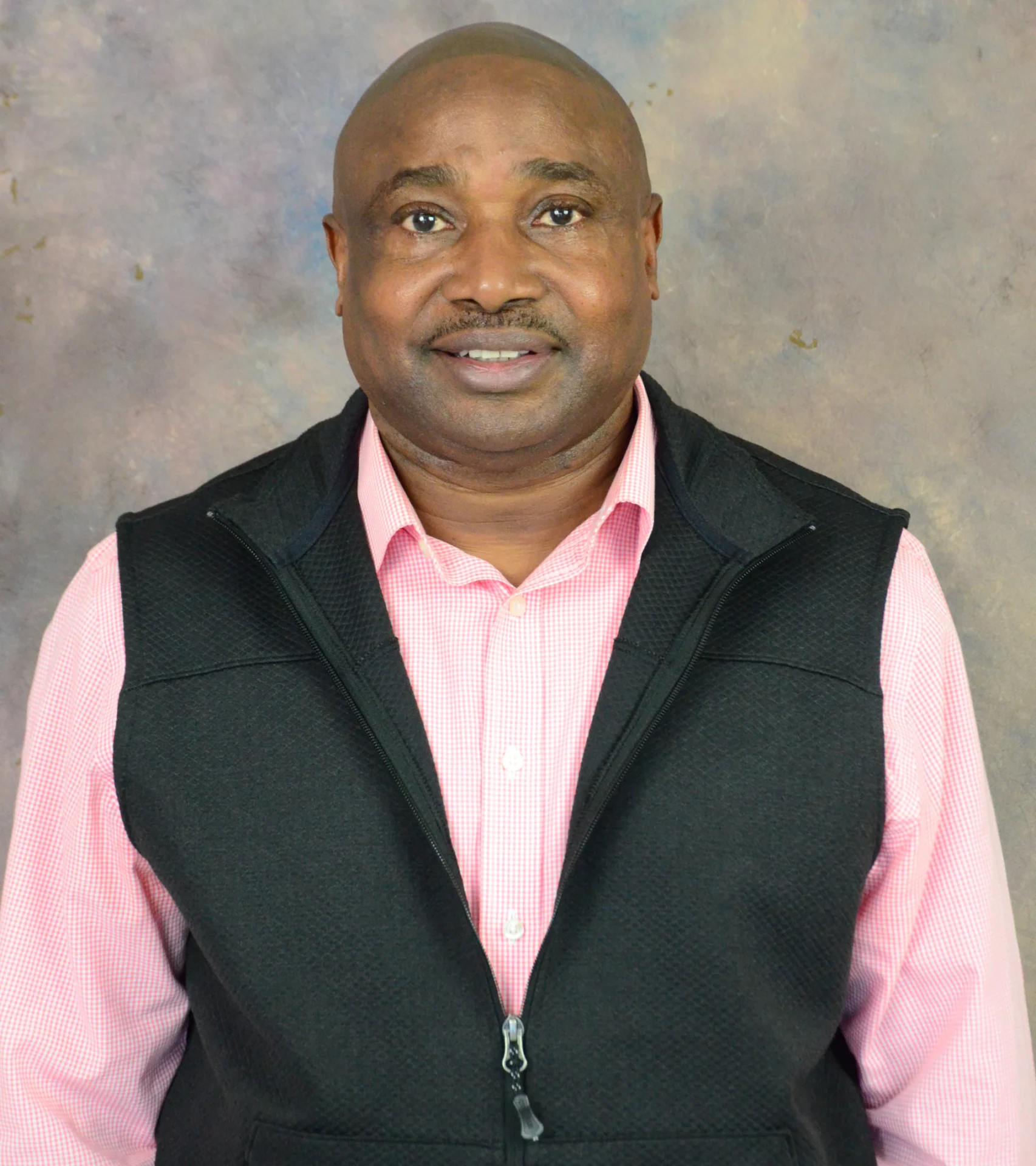 Smiling man in a pink shirt and black vest against a neutral background.